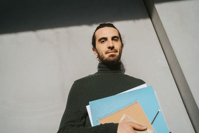 Low angle portrait of young male adult student holding book against wall at college on sunny day