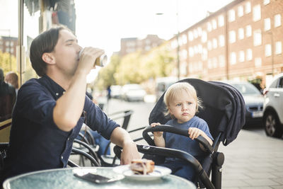 Baby boy looking away while sitting on stroller by father drinking at sidewalk cafe in city