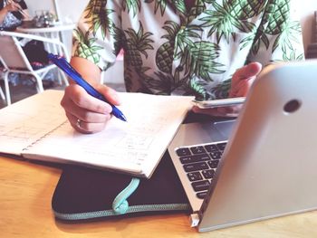 Midsection of woman using mobile phone while writing in book at table