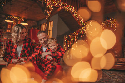 Smile active parents with small son in red checkered sleepwears waiting santa indoor.