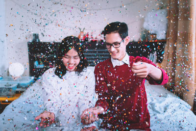 Cheerful couple sitting on bed with flying colorful confetti