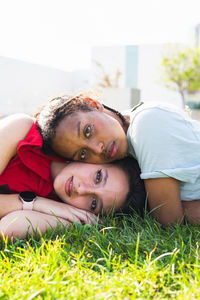 Charming multiracial female friends laying heads on each other and looking at camera while resting on grassy lawn in park