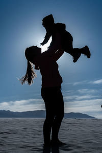 Rear view of man with arms outstretched standing at beach against sky during sunset