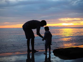 Side view of father and son on beach against sky during sunset