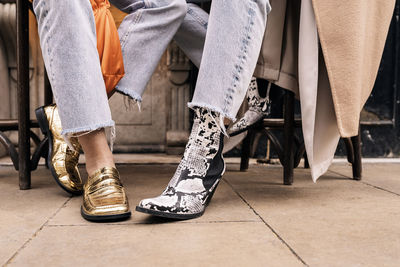 Legs of crop anonymous female friends in jeans and stylish footwear sitting together on chairs on street terrace in city