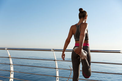 Rear view of woman exercising by railing while standing against sea