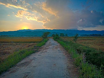 Dirt road amidst field against sky during sunset