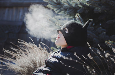Playful boy wearing knit hat while exhaling breath vapor at yard