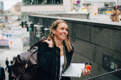 Mature businesswoman carrying luggage with laptop and coffee cup on staircase in city