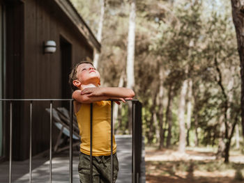 Little boy standing on veranda of modern cottage located in woods in summer