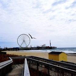 Ferris wheel by pier at sea against cloudy sky