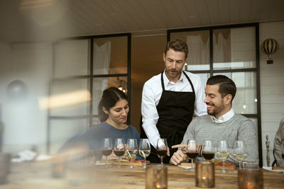 Man wearing apron standing by business people while pointing at wineglasses on table