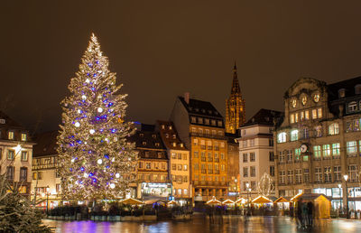 Illuminated christmas tree by buildings against sky at night