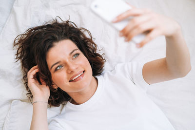 Young smiling brunette woman in home clothes using mobile on bed at home, top view