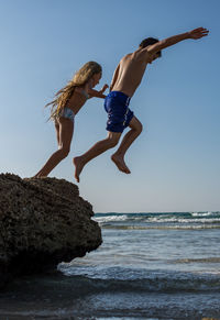 Siblings jumping from rock in sea against clear sky