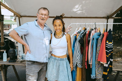 Senior man with hand on hip standing by female partner near clothes rack at flea market