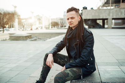 Portrait of hipster man sitting on footpath in city