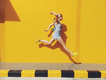 Side view of mid adult woman jumping on footpath against yellow wall