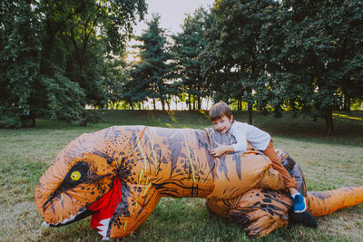 Portrait of boy sitting on person wearing dinosaur costume in park