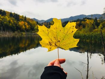 Close-up of hand holding autumn leaf against lake