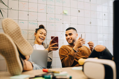 Smiling young businesswoman sitting with feet up on desk sharing smart phone with businessman against wall at creative o