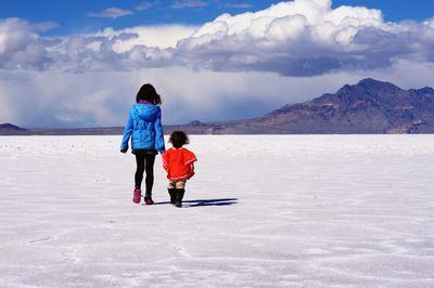 Rear view of sisters walking on snow covered field against cloudy sky 