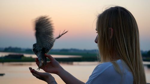 Woman holding bird against sky during sunset