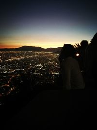 Rear view of people looking at illuminated cityscape against sky during sunset