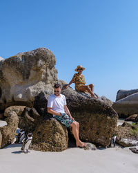 Rear view of woman sitting on rock against clear blue sky