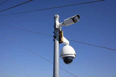 Low angle view of security camera by metallic cable against blue sky