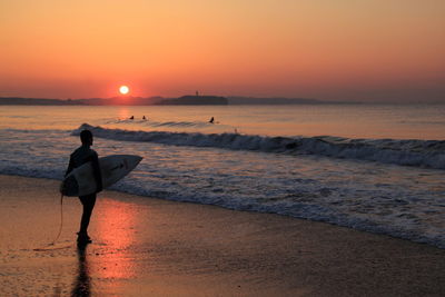 Man with surfboard standing on beach against sky during sunset
