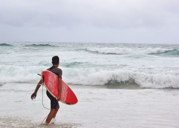 Rear view of man holding surfboard while walking in sea