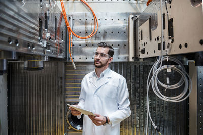 Man wearing lab coat and safety goggles at machine holding tablet