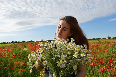 Beautiful young woman holding flowers in poppy field against sky