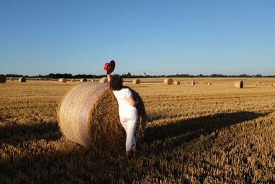 Rear view of woman leaning on hay bale at farm against clear sky