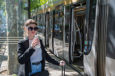 A business woman in sunglasses drinks water at a tram stop in the city, dressed