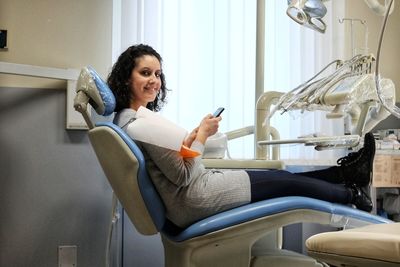 Portrait of smiling patient using mobile phone while sitting on chair in clinic