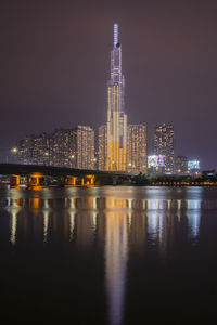 Illuminated buildings by river against sky at night