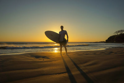 Man holding surfboard standing on beach against sky during sunset