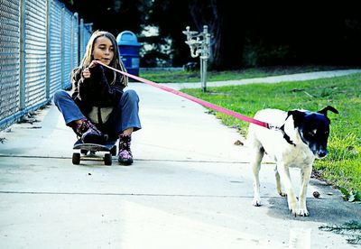 Girl sitting on skateboard while holding leash of dog at footpath