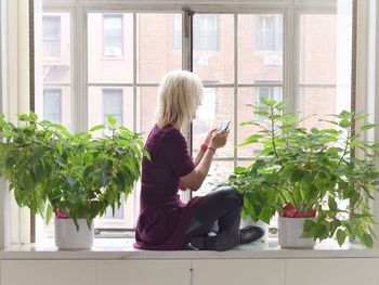 Side view of mature woman using mobile phone while sitting on window sill