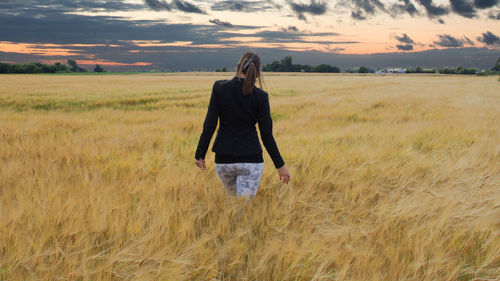 Woman walking on wheat field against sky during sunset
