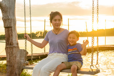 Smiling mother sitting on swing with son during sunset