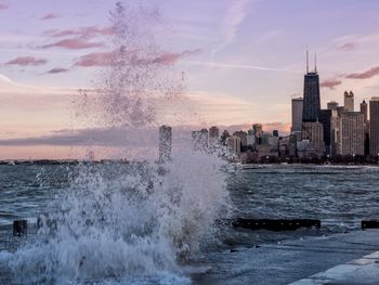 Water splashing in sea against sky at city during sunset