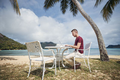 Young freelancer working on laptop from tropical destination. man sitting under palm trees on beach.