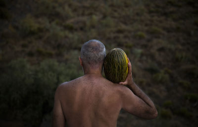 Rear view of shirtless man holding melon on shoulder in field