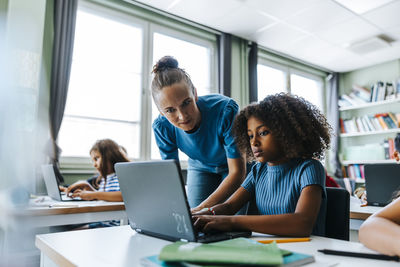 Female teacher assisting curly hair girl studying on laptop while sitting near desk in classroom at elementary school