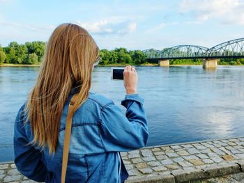Rear view of woman with long hair photographing river from mobile phone