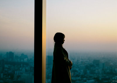 Woman standing by window during sunset