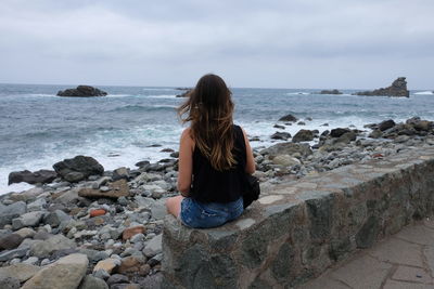 Rear view of woman sitting on rock at sea against sky 